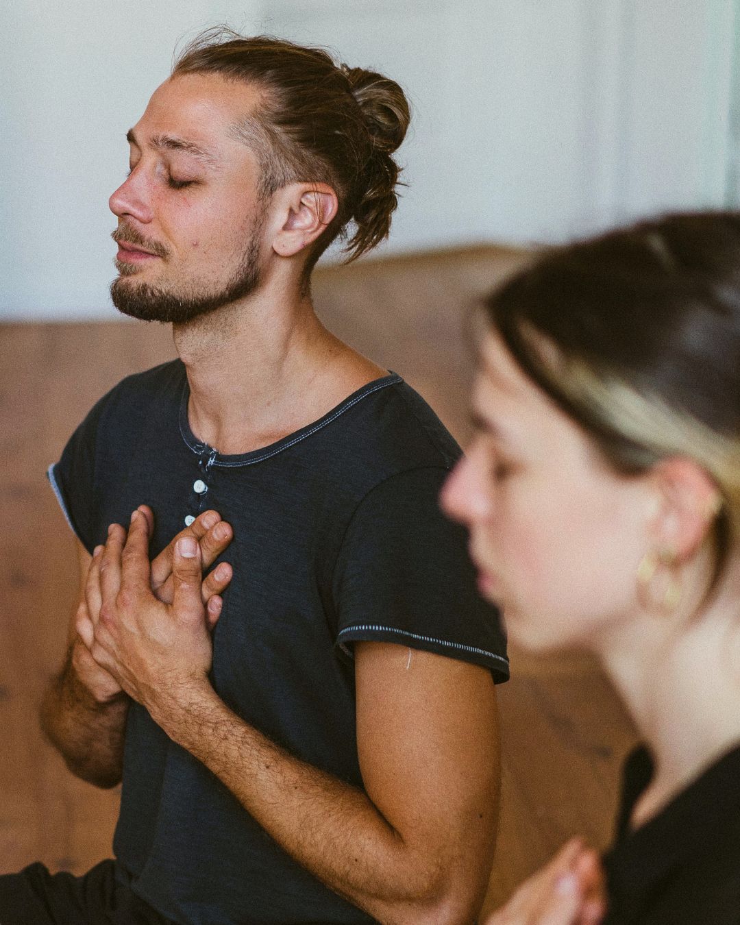 Teilnehmer sitzt mit geschlossenen Augen und Händen am Herzen in einer geführten Meditation, im Hintergrund weitere Person unscharf im Raum.